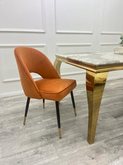 Rust velvet dining chair shown at an angle, with curved back and black, gold-tipped legs